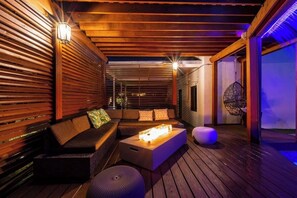 Eine Holzterrasse mit einer Sitzecke, bestehend aus einer Couch, Hocker und einem Tisch mit Lampe.