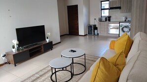 Ein modernes Wohnzimmer mit einem Flachbildfernseher, zwei kleinen Tischen, einer gelb gekissenen Couch, einer Küchenzeile mit Geräten und einer Tür zu einem weiteren Raum.