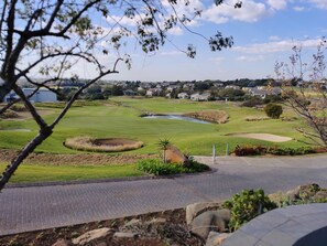 Um campo de golfe com lago, bunkers de areia e vista para uma área residencial ao fundo.