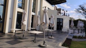 Ein Außenbereich mit Tischen und Sonnenschirmen vor einem Gebäude mit großen Fenstern und einer überdachten Terrasse.