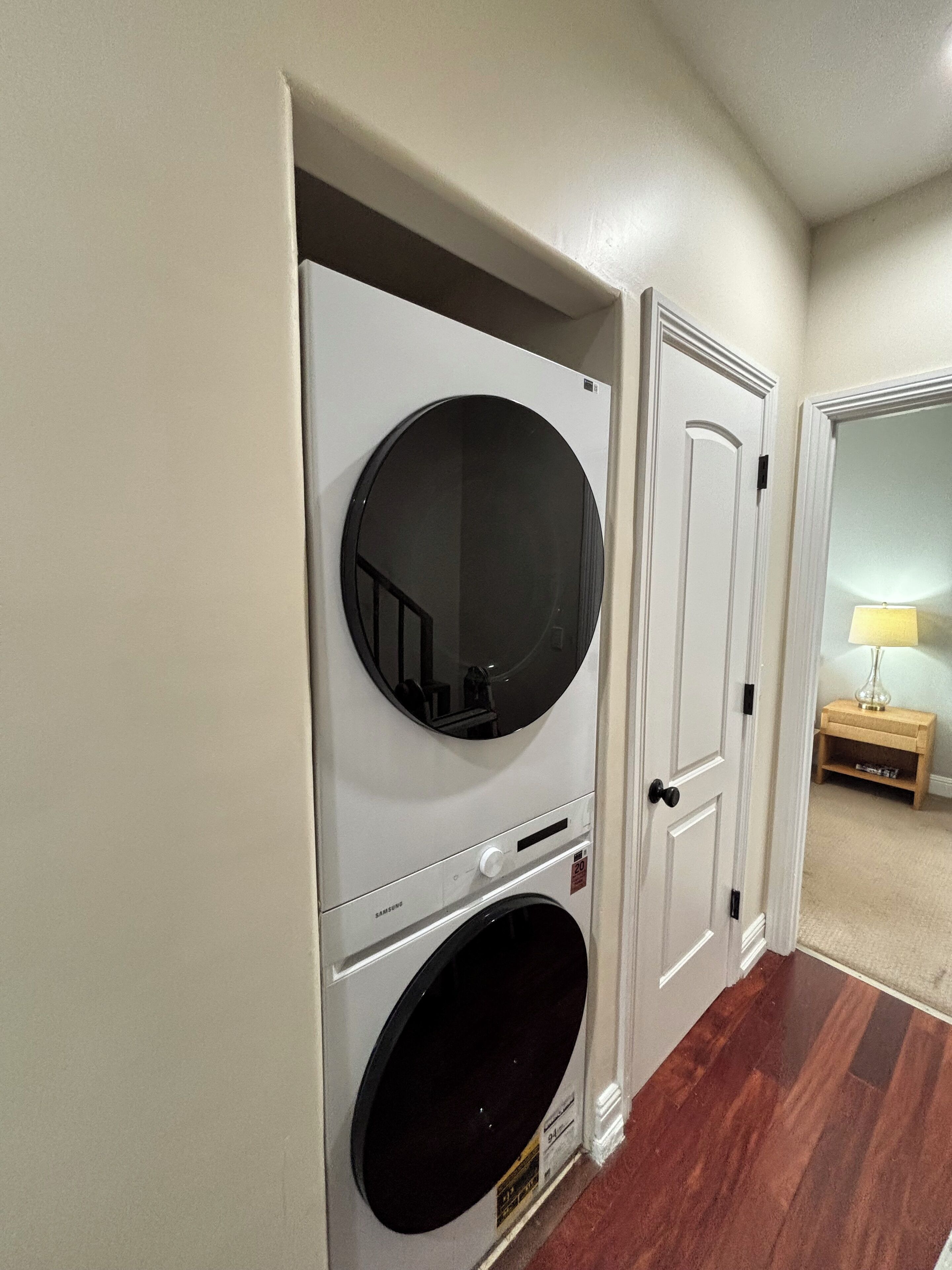 A laundry room with a front-loading washer and dryer, a door, and a small wooden table with a lamp.