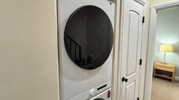 A laundry room with a front-loading washer and dryer, a door, and a small wooden table with a lamp.