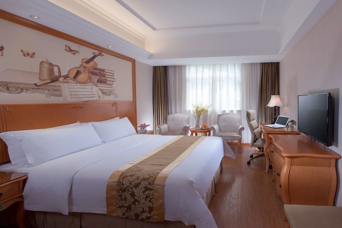 Ein Hotelzimmer mit einem großen Bett, einem Schreibtisch mit Fernseher, einem Sessel und einem Fenster mit Vorhängen.