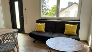 A room with a black sofa, two yellow cushions, a round table with a glass top, and a wooden chair with a metal frame.