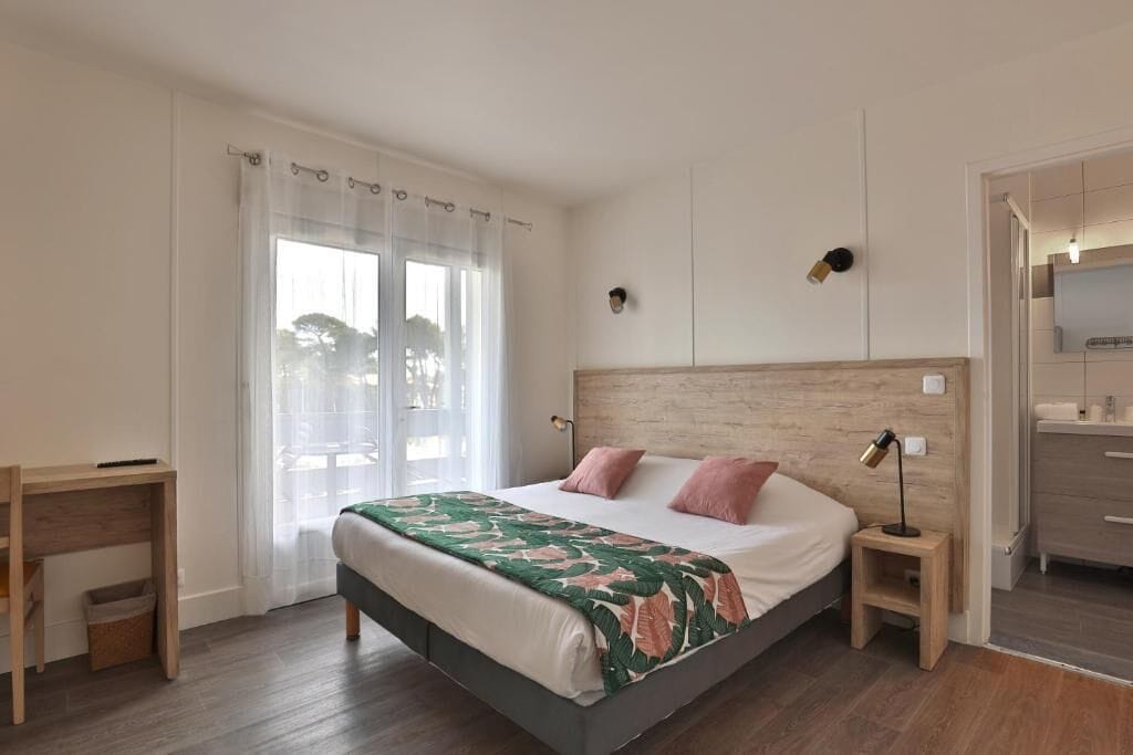 Un dormitorio con cama, mesitas de noche, escritorio, espejo y baño visible a través de una puerta abierta.