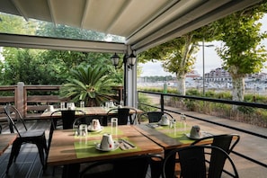 An outdoor dining area with tables set for two, surrounded by greenery and overlooking a marina with boats.