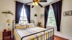 Un dormitorio con cama, ventilador de techo, mesita de noche, lámpara, ventana con cortinas y una planta en la pared.