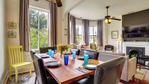 Comedor con mesa de madera dispuesta para cuatro personas, rodeada de sillas, y una televisión y chimenea al fondo.