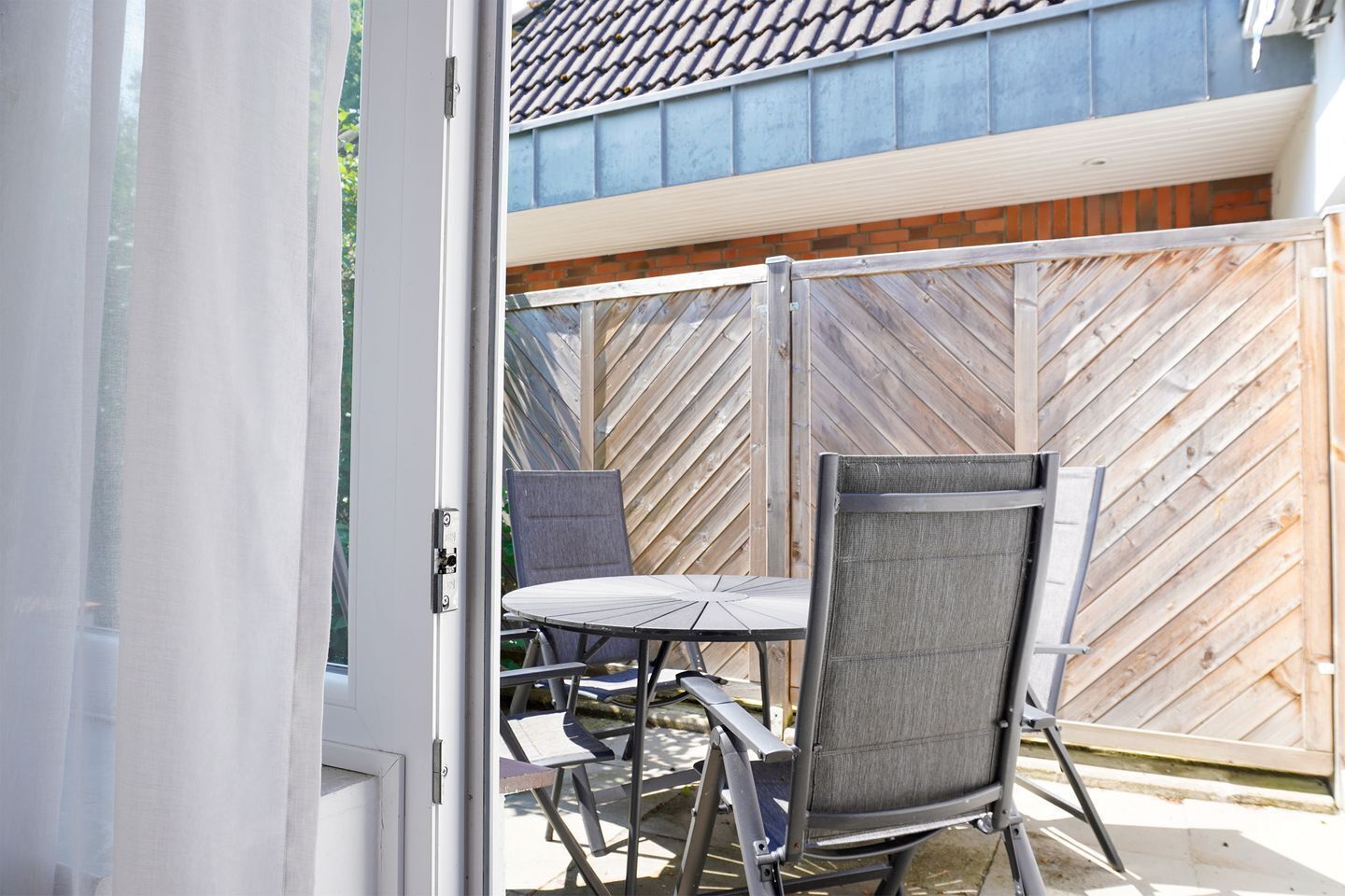 Eine geöffnete Tür, die auf eine Terrasse mit Tisch und Stühlen führt, ein Holzzaun und im Hintergrund ein Gebäude mit einem Ziegeldach.