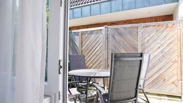 Eine geöffnete Tür, die auf eine Terrasse mit Tisch und Stühlen führt, ein Holzzaun und im Hintergrund ein Gebäude mit einem Ziegeldach.