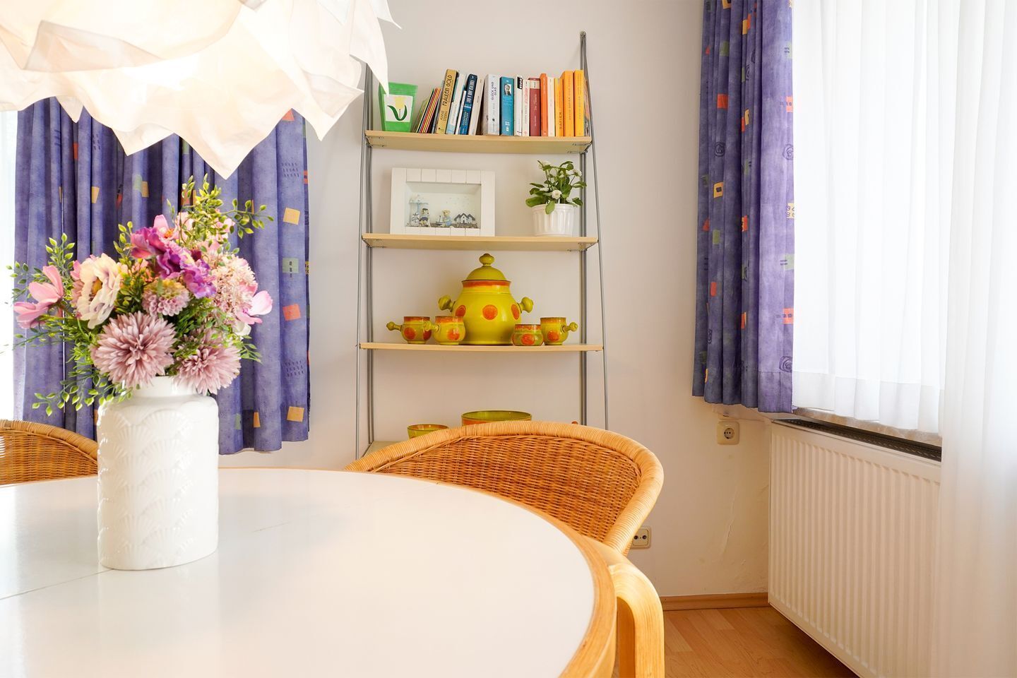 A room with a round table, wicker chairs, a white vase with flowers, a bookshelf with books and decorative items, and purple curtains.