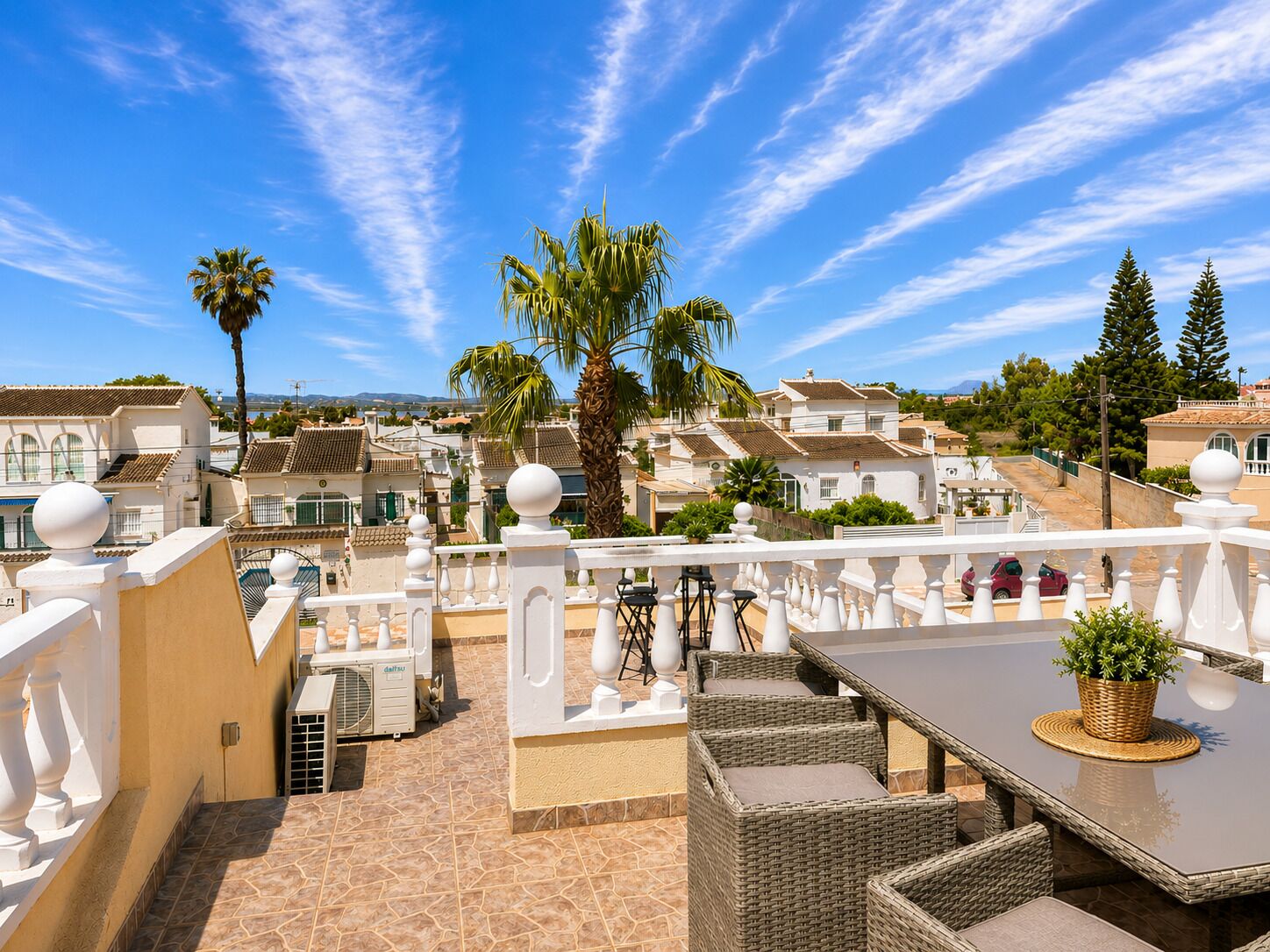 A terrace with outdoor furniture, a white balustrade, and a view of a residential area with houses and palm trees.