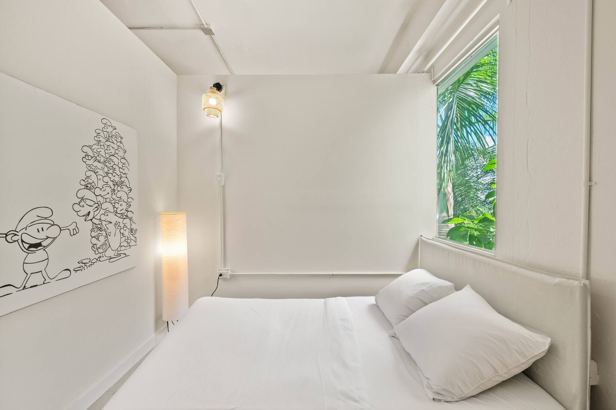 A bedroom with a bed, a wall-mounted light, a window with a view of greenery, and a wall art featuring cartoon characters.