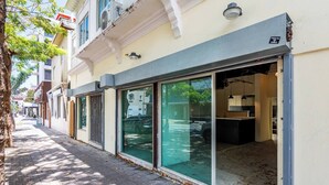 Um edifício com grandes janelas de vidro, interior visível com balcão e luminárias pendentes, e uma rua arborizada.