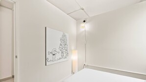 Quarto minimalista com uma cama branca, uma obra de arte na parede e um abajur.