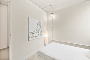 Ein schlicht eingerichtetes Schlafzimmer mit einem weißen Bett, einem Wandbild und einer Nachttischlampe.