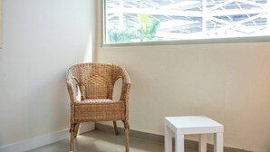 Uma cadeira de vime ao lado de uma mesa branca num quarto com uma janela grande.
