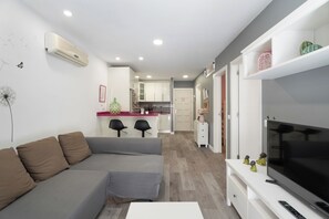 A modern living room with a gray sectional sofa, a flat-screen TV on a white stand, and a kitchen in the background. - Charming 2-Bedroom Apartment in Puerto de Santiago (Puerto de Santiago)