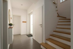 A hallway with a wooden staircase, a door with a handle, a small plant on a cabinet, and a light switch on the wall. - Villa Lykka (100631-K1) - Kastelir (Kastelir)