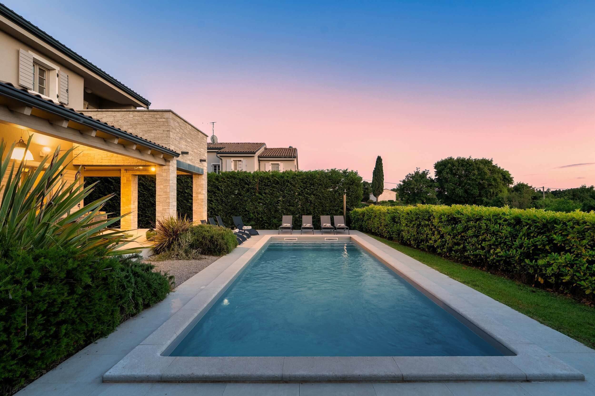 A rectangular swimming pool with a stone border, surrounded by a well-maintained hedge and a row of lounge chairs.