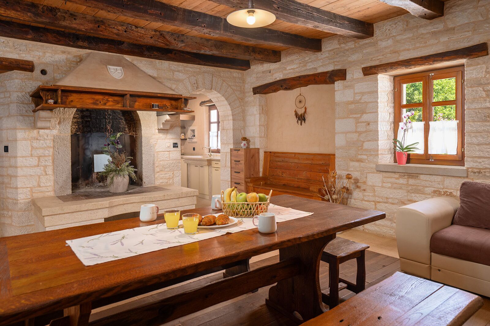 Une cuisine rustique aux murs de pierre, une table en bois où est servi le petit-déjeuner et un coin salon confortable.