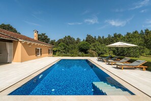 En rektangulær swimmingpool med fliseterrasse, et loungeområde med liggestole og parasol samt en bygning med flisebelagt tag og beige vægge.