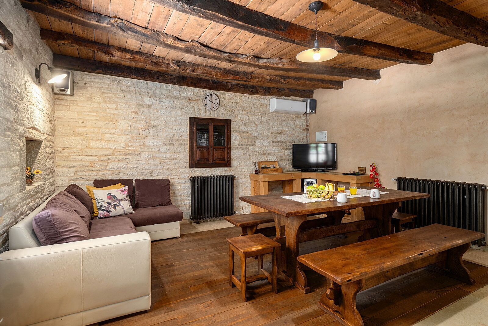 Una habitación rústica con pared de piedra, vigas de madera en el techo, un sofá, una mesa con bancos, un televisor y un radiador.