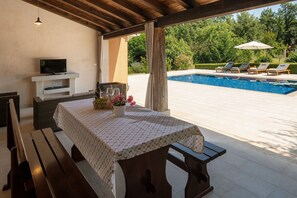 Eine Terrasse im Freien mit Esstisch, Fernseher und Poolbereich mit Liegestühlen.