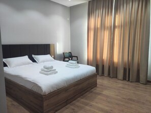 Cama bem arrumada com roupa de cama e toalhas brancas, cabeceira de madeira e uma cadeira em quarto com piso de madeira e cortinas.