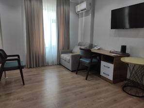 Quarto moderno com piso de madeira, sofá cinza, escrivaninha com cadeira, TV fixada na parede e ar condicionado.