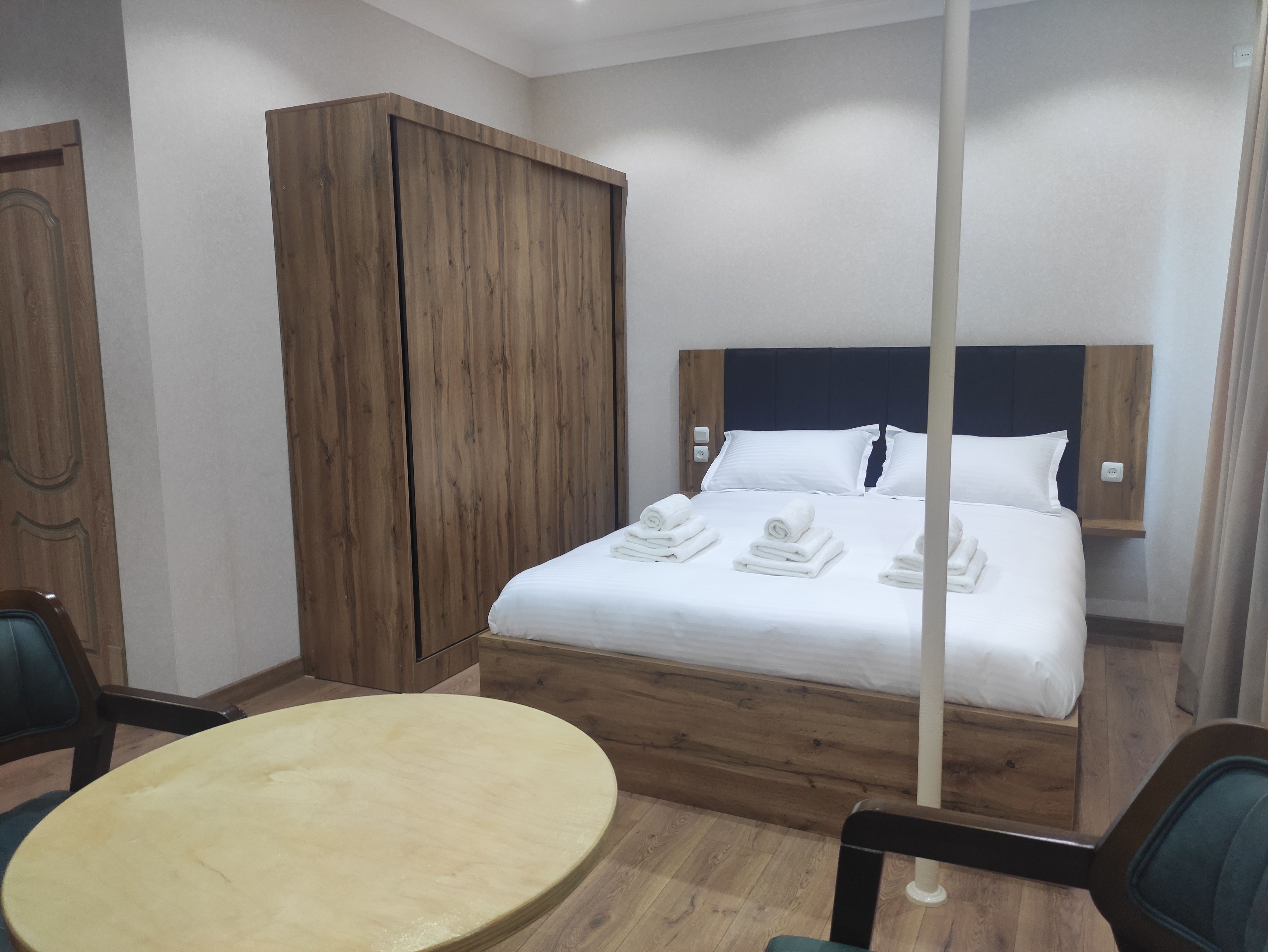 A hotel room with a bed, a wooden wardrobe, a round table, and a chair.