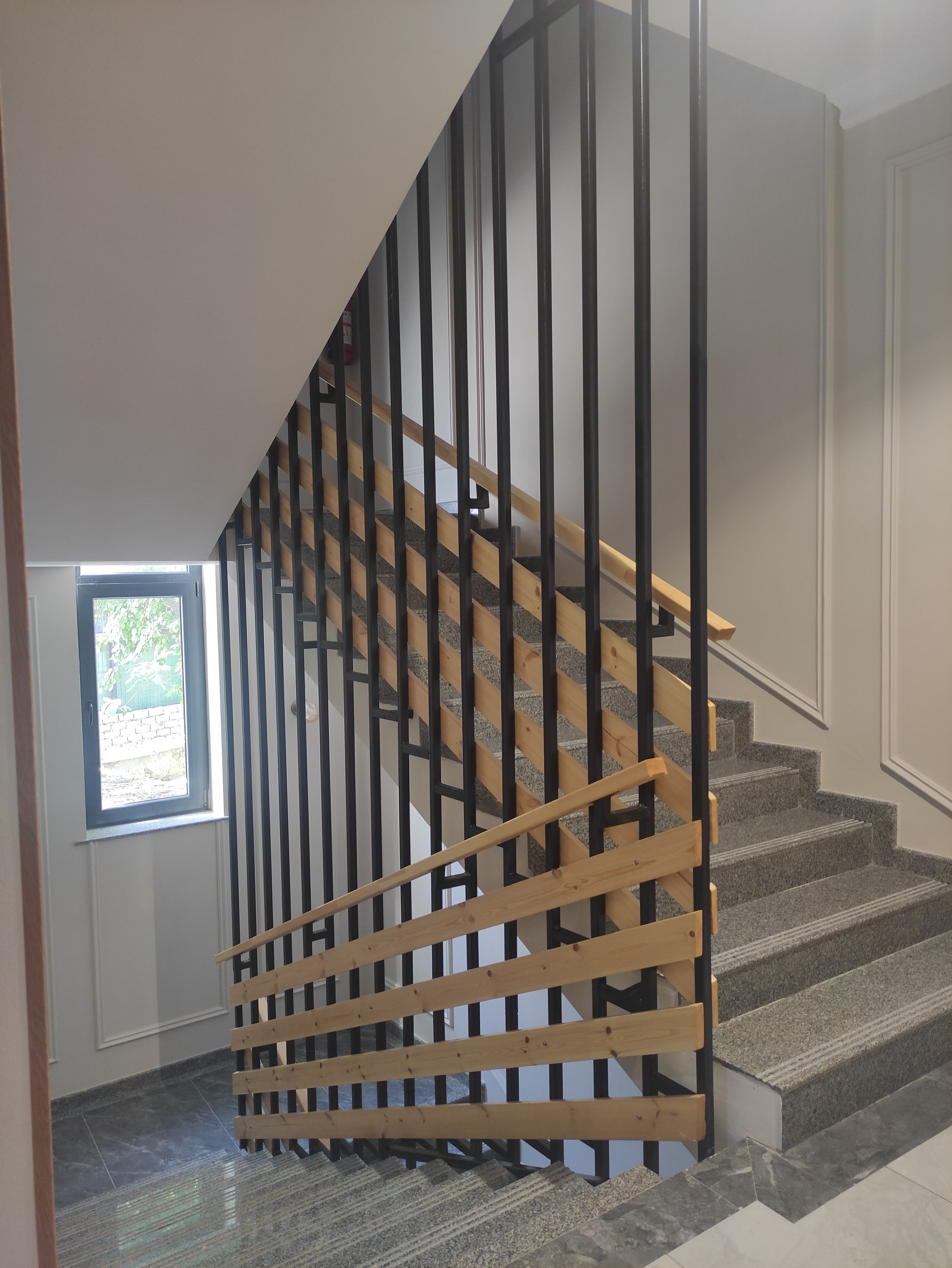 Una escalera con barandilla de madera y balaustradas negras, que lleva a un rellano con una puerta y un ventanal.