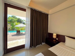 Habitación de hotel con cama, mesita de noche, lámpara, ventana con cortinas y vistas a una piscina al aire libre.