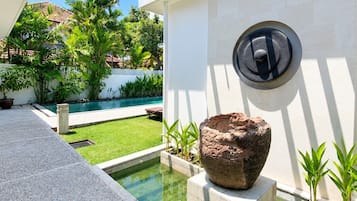 Zona de piscina con una escultura de piedra, una pared blanca y una pérgola.