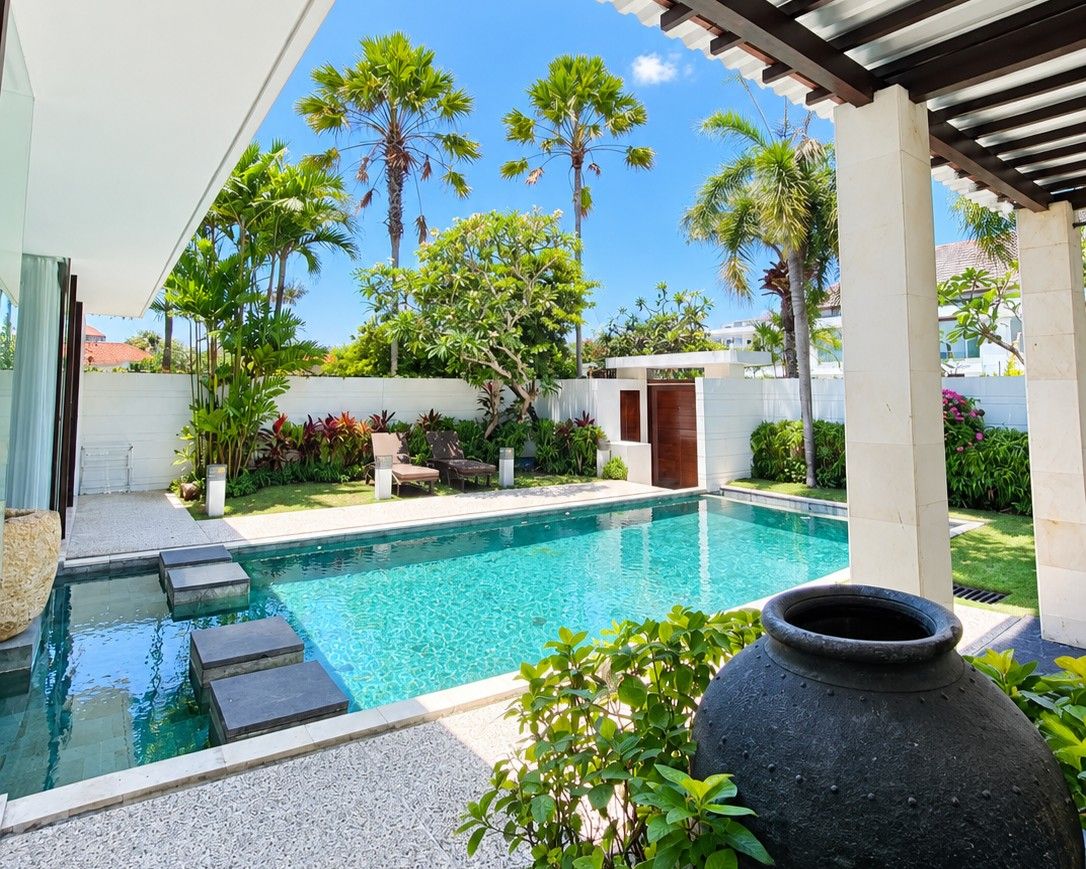 Una piscina con una pérgola, rodeada de exuberante vegetación y un cielo azul despejado.
