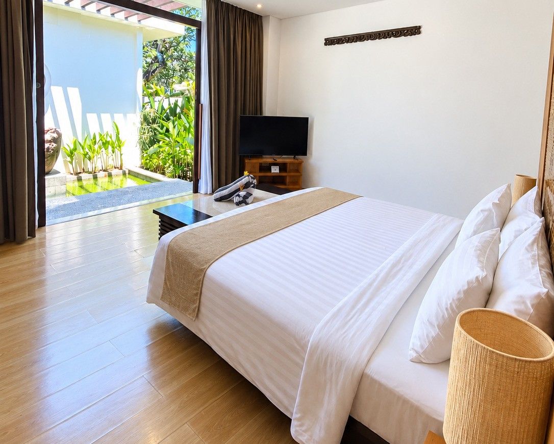 A hotel room with a bed, a television, a small table, and a view of a garden through a glass door.