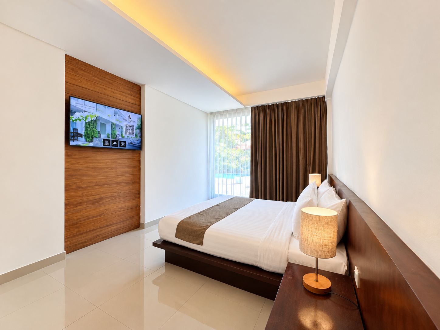 A modern hotel room with a bed, a television mounted on the wall, a bedside table with a lamp, and a view of the outdoors through the window.
