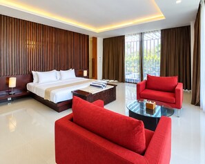 A modern hotel room with a large bed, a red sofa, a glass coffee table, and a view of greenery outside. - The Wangsa Hotel and Villas (Nusa Dua)