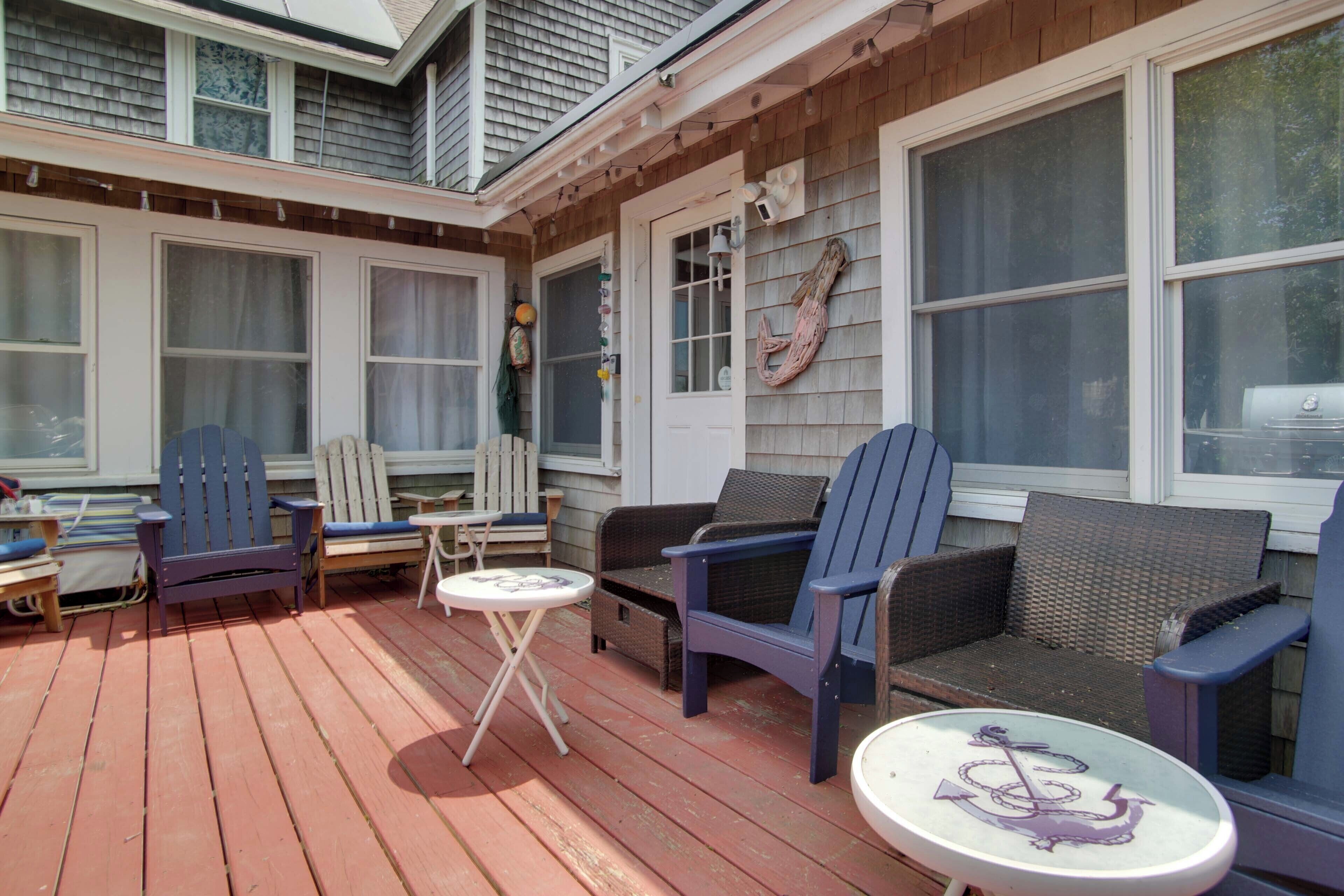 A covered outdoor deck with wooden flooring, two blue Adirondack chairs, a wicker sofa, and a small round table with a decorative anchor.