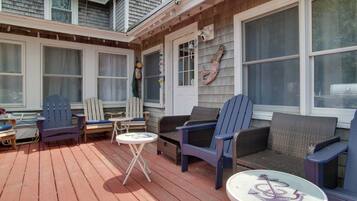 A covered outdoor deck with wooden flooring, two blue Adirondack chairs, a wicker sofa, and a small round table with a decorative anchor.