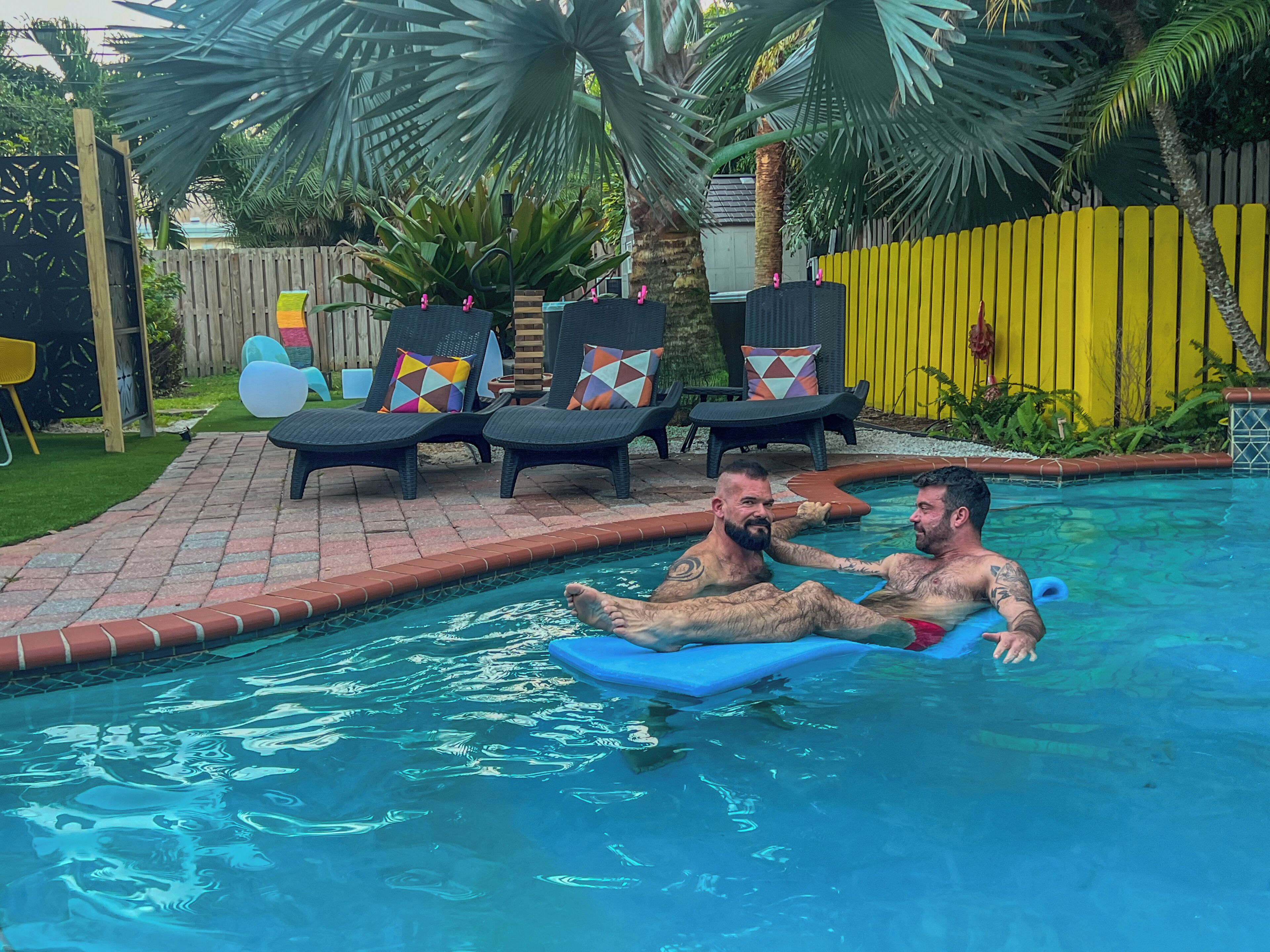 A swimming pool with two people on a blue float, surrounded by lounge chairs with colorful cushions, a wooden fence, and palm trees.