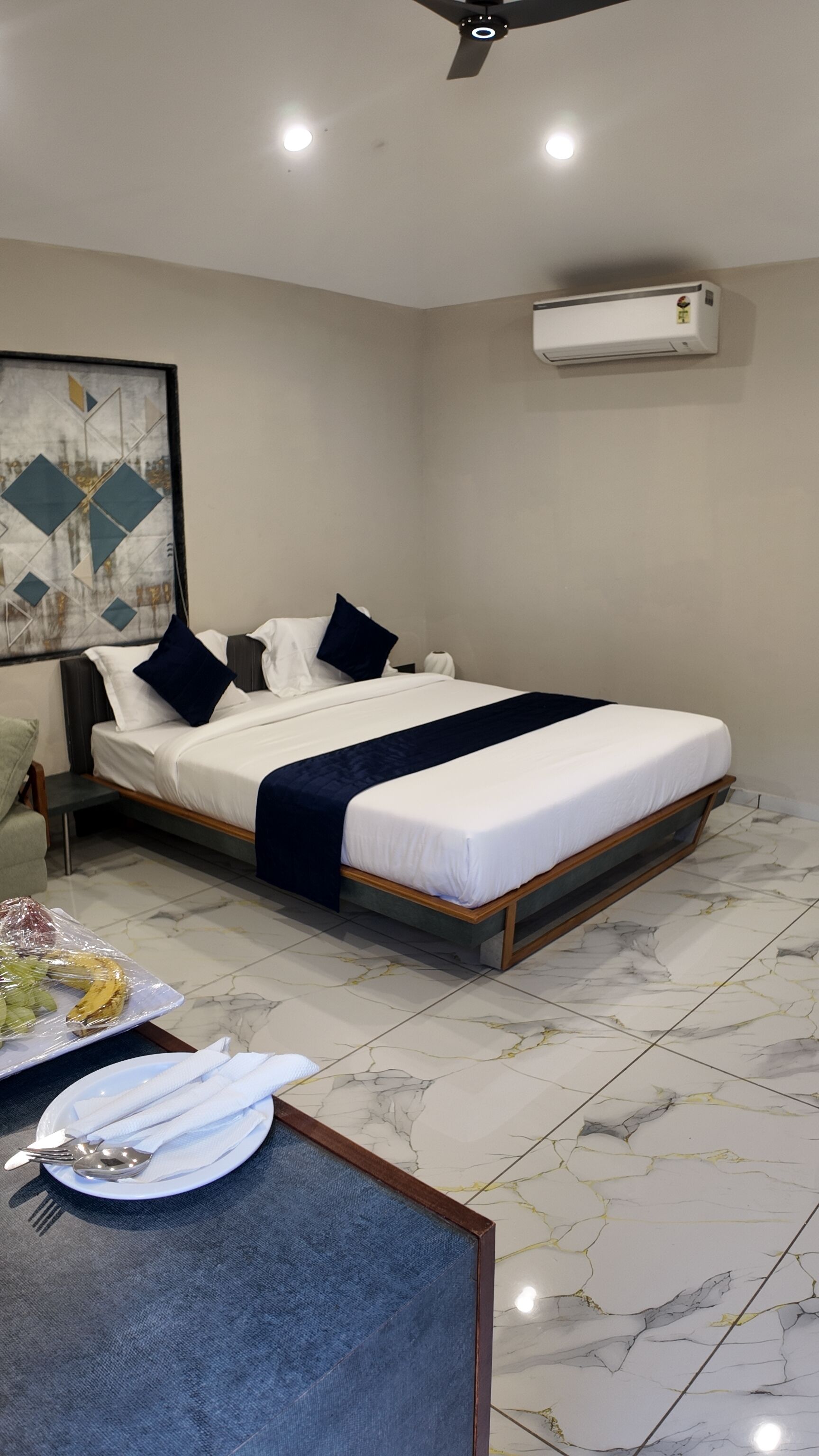 A hotel room with a bed, a ceiling fan, an air conditioning unit, a dining table with a plate and utensils, and a wall with a geometric painting.