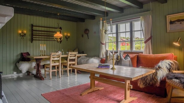 A cozy room with a wooden table, chairs, a sofa, a window with curtains, and a hanging lamp.