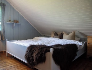 A bedroom with a sloped ceiling, a bed with a white mattress and pillows, a wooden nightstand, and a fur throw on the bed.