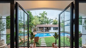 A view through glass doors to a backyard with a swimming pool, a covered patio, and potted plants. - Gorgeous Grand Pool Villa in D2 by Ray (Ho Chi Minh City)
