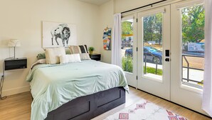 Ein Schlafzimmer mit Bett, Nachttisch, Lampe, Fenster mit Ausblick und einer Tür.