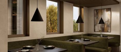 A modern dining area with green upholstered seating, pendant lighting, and large windows offering views of trees.