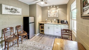 Une cuisine compacte équipée d’un réfrigérateur, d’un four à micro-ondes et de meubles blancs. On y trouve également un ventilateur de plafond, un tapis et deux chaises en bois.