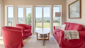Una habitación con un sofá rojo, un sillón rojo, una mesita redonda y grandes ventanales con vistas al paisaje.