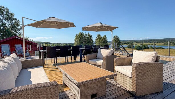 Terraza exterior con mobiliario de mimbre, dos sombrillas grandes y vistas a un lago y colinas.